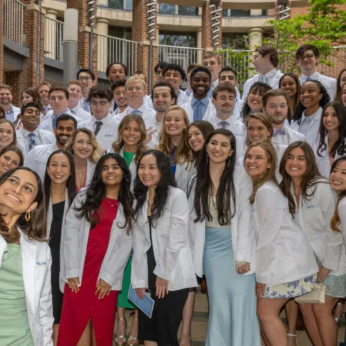 One person takes a selfie of a large group of students in white coats.