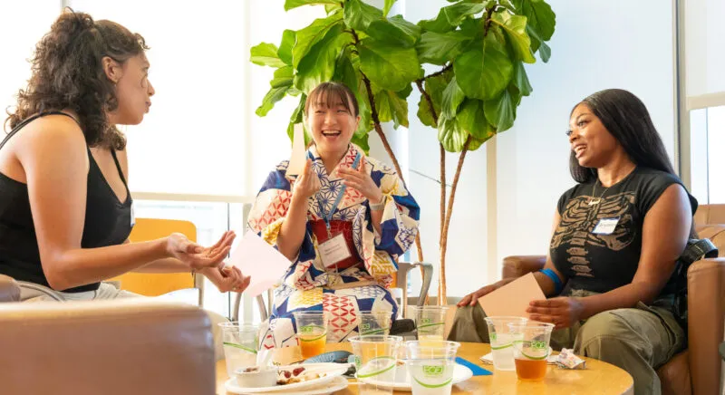 Three UNC students sit in cushioned chairs around a wooden circular table talking and laughing.