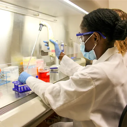 Female student wearing laboratory coat, mask, and goggles using pipette.